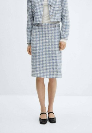 Femme portant un tailleur à carreaux bleu et blanc avec des chaussures Mary Jane noires, se tenant devant un fond blanc uni.