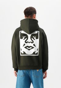 Olijfgroene hoodie met een grote witte grafische afbeelding van een gezicht op de achterkant, voorzien van een trekkoordkap en geribde manchetten. Blauwe jeans zijn zichtbaar eronder.