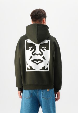 Olivgrüner Hoodie mit einem großen weißen Gesichtsdruck auf dem Rücken, ausgestattet mit einer Kordelzugkapuze und gerippten Bündchen. Blaue Jeans sind darunter sichtbar.