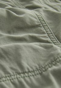 Textiel in een groene kleur met structuur en zichtbare stiksels, dat zachte, gekreukte gedeeltes en een gladde oppervlakte toont. Het materiaal lijkt lichtgewicht en ademend te zijn.