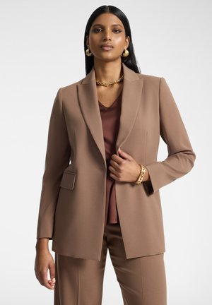 Mujer con traje marrón, pendientes de aro dorados, collar y pulsera, de pie frente a un fondo blanco liso.