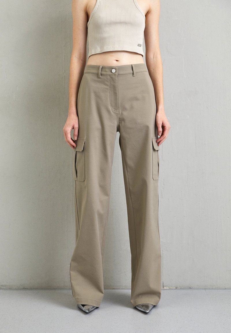 Personne portant un pantalon cargo beige taille haute avec des poches latérales et un débardeur beige côtelé court, debout contre un mur uni.