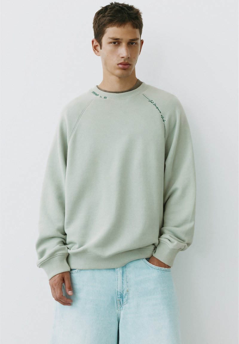 Jeune homme portant un sweat-shirt gris clair et un jean bleu clair, debout devant un fond blanc avec une expression neutre.