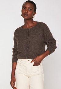 Frau trägt eine dunkelbraune, geknöpfte Strickjacke mit langen Ärmeln und cremefarbene, hochtaillierte Hose und posiert mit einer Hand in der Tasche.