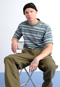 Črtasta majica v zelenih in modrih odtenkih, iz bombažne mešanice, kratki rokavi, okrogla izreza. Oljčno zeleni cargo hlače. Srebrni prstani in tetovaža na roki.