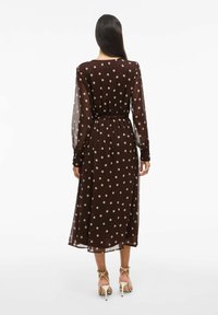 Bruine jurk met polka dots, lange doorschijnende mouwen, een ceintuur op taillehoogte en een lengte tot onder de knie. Lichtgewicht stof met een gladde textuur.