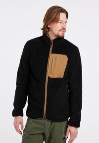 Zwarte fleece jas met een zachte textuur, ritssluiting, hoge kraag en een beige borstzak. Gecombineerd met groene broek.