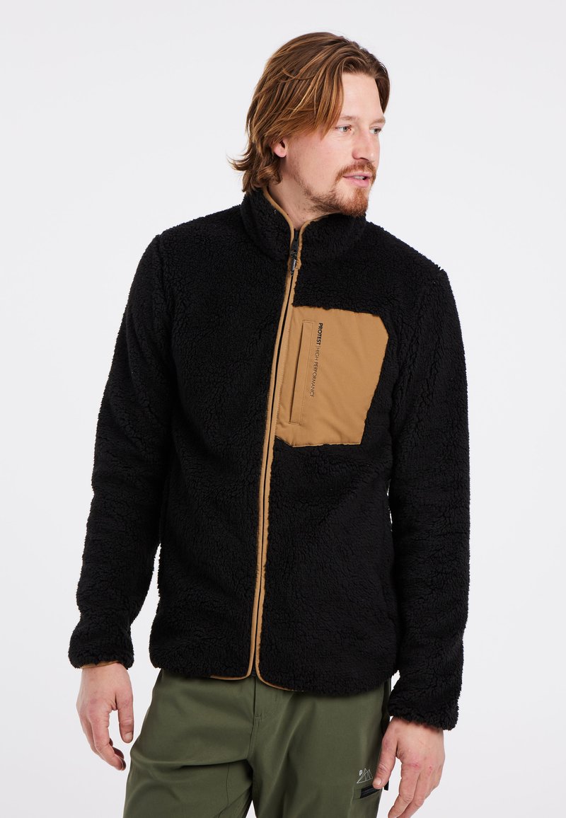 Zwarte fleece jas met een zachte textuur, ritssluiting, hoge kraag en een beige borstzak. Gecombineerd met groene broek.