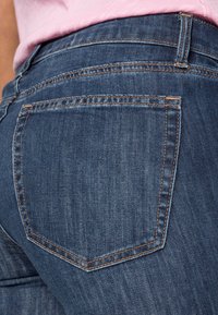 Jeans de mezclilla azul oscuro con un bolsillo trasero que presenta costuras en contraste de color naranja. La tela tiene una textura suave y un leve desgastado.