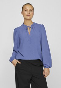 Blusa di un leggero viola con maniche lunghe gather ed uno scollo a chiave rifinito con un fiocco. Texture liscia abbinata a pantaloni scuri.