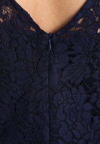 Primo piano di tessuto in pizzo blu scuro con motivi floreali e una cerniera centrale su uno sfondo di pelle chiara.