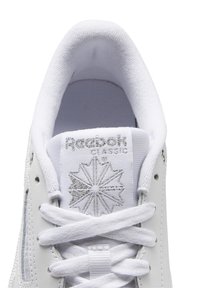 Gros plan sur la languette d'une basket blanche Reebok Classic avec un logo argenté et des lacets blancs noués en motif croisé classique.