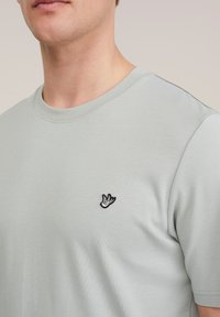 Man met een lichtgrijs T-shirt met een klein zwart-wit geborduurd vogel-logo op de linkerborst, gefotografeerd vanaf de nek en schouder.