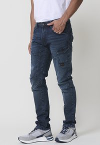 Jeans de denim azul oscuro con un corte ajustado, que cuentan con un bolsillo tipo cargo en el muslo, costuras detalladas y botones de metal. Combinados con zapatillas grises.