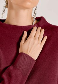 Close-up van een hand van een persoon die rust op een bordeauxrood sweater nabij de kraag, met hangende kristallen oorbellen zichtbaar tegen een lichte achtergrond.