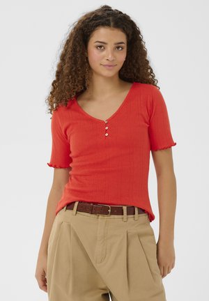 Jeune femme aux cheveux bouclés portant un haut rouge à manches courtes avec des boutons et un pantalon plissé beige avec une ceinture marron, debout devant un fond blanc.