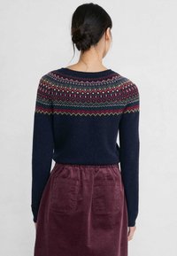 Pull en maille bleu marine avec un empiècement jacquard arborant des motifs rouges, verts et blancs, associé à une jupe en velours côtelé bordeaux avec deux poches arrière.