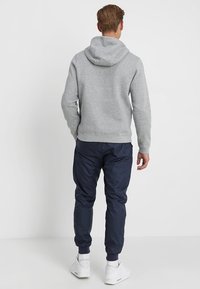 Grå sweatshirt med huva, kombinerat med marinblå joggingbyxor. Har ribbade muddar och midja. Skorna är vita sportskor.