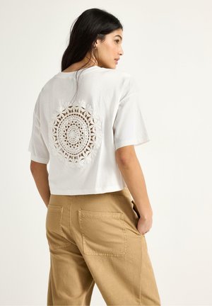 Pitkähiuksinen nainen, jolla on valkoinen lyhyt T-paita, jossa on pyöreä virkkauskuvio selässä, ja tan-väriset korkeavyötäröiset housut.