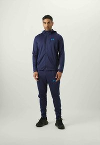 Muškarac stoji u tamnoplavom trening odijelu Under Armour s duksericom na patentni zatvarač i odgovarajućim hlačama, uparenim s crnim tenisicama na jednobojnoj pozadini.