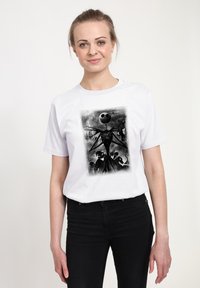 Bílé tričko s černobílým grafickým potiskem stylizované postavy s lebkovitou tváří uprostřed temné, dramatické scény.