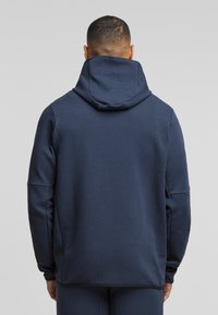 Tamno plavi dukserica napravljena od mekane tkanine, s velikom kapuljačom, dugim rukavima i opuštenim krojem. Dizajn na leđima je jednostavan bez vidljivih logotipa.