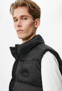 Schwarze Steppweste mit hochgeschlossenem Kragen, Reißverschluss und Logopatch. Hergestellt aus glänzendem, gestepptem Material, getragen über einem weißen Hemd.