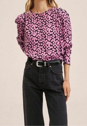 Vrouw die een roze en zwarte blouse met lange mouwen en polkadot draagt, ingestopt in een zwarte, hooggehate spijkerbroek met een zwarte riem.