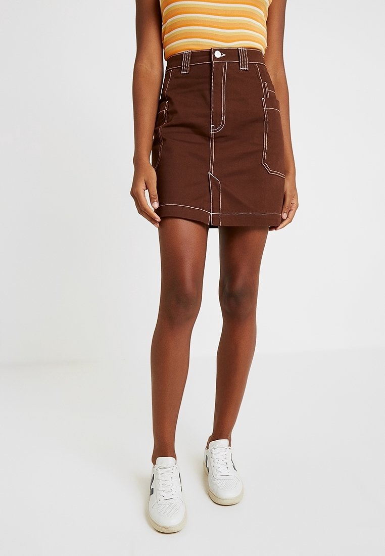 Femme portant une minijupe marron avec des coutures blanches, un haut rayé jaune et des baskets blanches, debout devant un fond blanc uni.