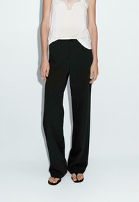 Pantalons larges noirs en tissu lisse, avec des poches devant et une coupe décontractée, associés à un débardeur blanc simple.
