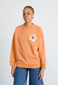 Sweat-shirt orange avec un grand motif de fleur d'hibiscus blanc sur la poitrine gauche. Fabriqué à partir d'un matériau doux, il présente des poignets et un ourlet côtelés.