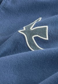 Tissu bleu avec broderie texturée d’un oiseau stylisé délimité par un fil blanc.
