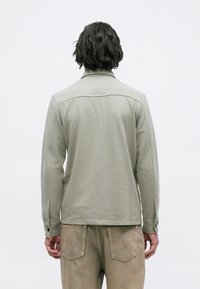 Άτομο με σγουρά σκούρα μαλλιά που φοράει ανοιχτό γκρι μακρυμάνικο μπλουζάκι και ξεθωριασμένα μπεζ τζιν, φαίνεται από πίσω με φόντο απλή χρώμα.