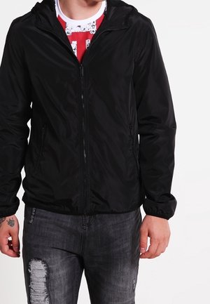 Homme portant une veste noire zippée par-dessus une chemise rouge et blanche avec un jean noir déchiré, debout devant un fond uni.