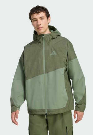 Grüne Windjacke mit Kapuze, kontrastierenden Texturen, einem Gittermuster im oberen Bereich und Reißverschlusstaschen.