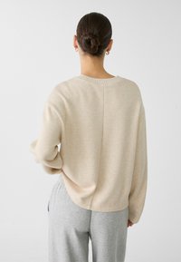 Beige Strickpullover mit Rundhalsausschnitt, langen Ärmeln, überschnittenen Schultern und lockerer Passform, kombiniert mit hellgrauen Jogginghosen.
