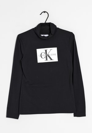 Czarna koszulka z długim rękawem i golfem, z białym prostokątnym logo z napisem "cK CALVIN KLEIN JEANS" na piersi, wisząca na czarnym wieszaku.