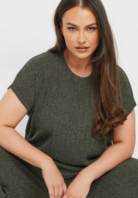 Tumma vihreä neulepaita lyhyillä hihoilla, pyöreällä pääntiellä ja ribatulla pinnalla. Hiukset ovat vapaana, istuu kädet reisillä.