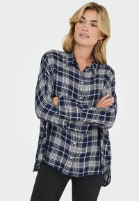 Plava žena s opuštenom kosom nosi srebrne naušnice u obliku prstena i košulju karo uzorka u tamnoplavoj i bijeloj boji, s prekriženim rukama, gleda u stranu.