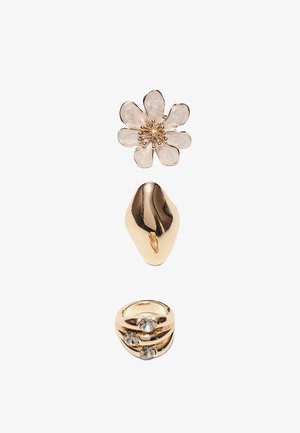 Trois bagues en or : la première ornée d'une fleur en émail blanc, la deuxième présentant une forme irrégulière lisse, la troisième décorée de trois pierres précieuses transparentes.