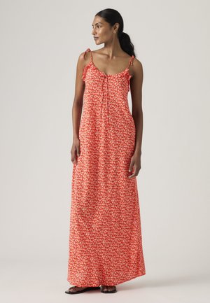 Mujer de cabello oscuro con un vestido largo rojo sin mangas con estampado floral y tirantes con lazos, de pie y mirando hacia un lado contra un fondo liso.