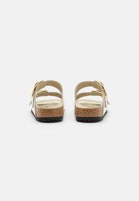 Witte open teen sandalen met een kurken voetbed, voorzien van een platte zwarte zool en een decoratieve strikaccent op de band. Achteraanzicht weergegeven.