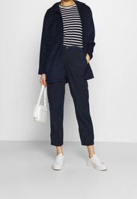 Giacca di velluto a coste blu navy sopra una camicia a righe bianca e blu navy, abbinata a pantaloni cargo blu navy e sneakers bianche, mentre tiene una borsa a tracolla bianca.