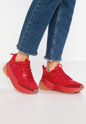 Osoba nosząca czerwone masywne sneakersy i niebieskie skrócone dżinsy z postrzępionym dołem stojąca na białej podłodze.