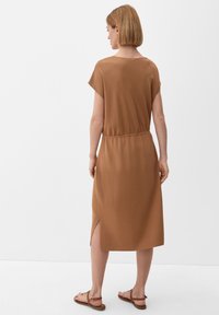 Bruine, korte jurk met een losse pasvorm, geaccentueerde taille, zijsplitten en een gladde textuur, gecombineerd met platte sandalen.