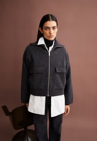 Donkergrijze wollen zip-up jas met twee voorzakken, geribbelde zoom en hoge kraag, gedragen over een wit shirt en donkere broek.