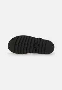 Black rubber sandal sole with textured ridges and a rectangular logo in the center. Thick, durable design with a non-slip surface.