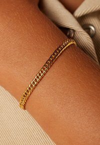 Gouden kettingarmband op een huidkleurige pols, met in elkaar grijpende schakels met een gladde, glanzende afwerking en een subtiele textuur.