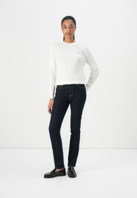 Hvit strikket genser med teksturert mønster, kombinert med mørkeblå bukser og svarte loafers. Modellen står mot en hvit bakgrunn.