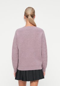 Pulôver roxo de malha com padrão de trancinhas nas mangas, gola redonda e bainha canelada, combinado com uma saia preta plissada.